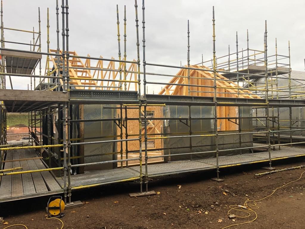 Timber roof trusses erected over a sheathed and membrane-wrapped timber frame, seen through the scaffold